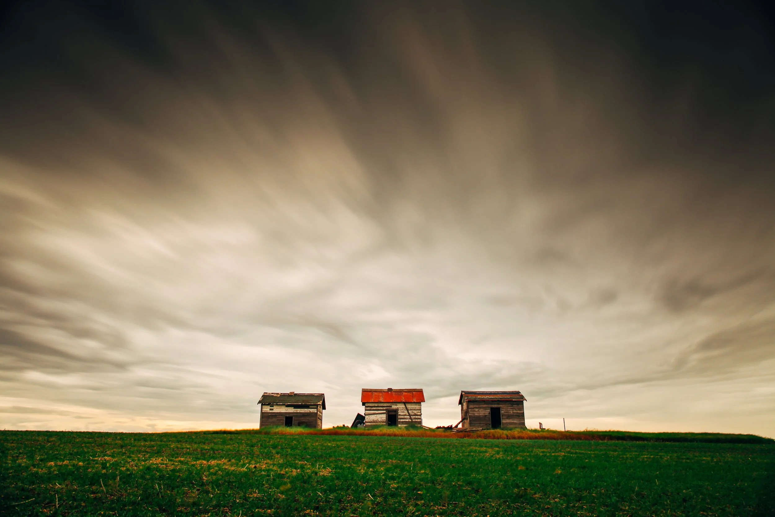Three old, weathered wooden houses on a grassy hill with a dramatic cloudy sky above.