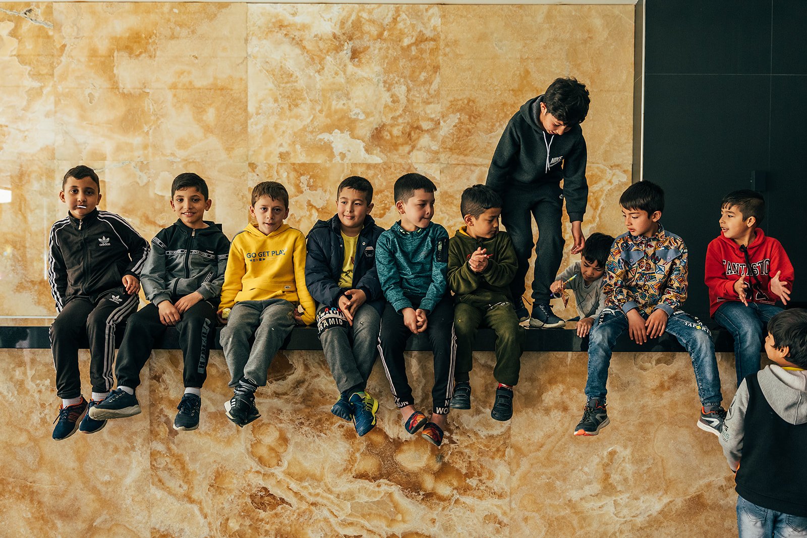 Group of children sitting on a ledge with one standing, in front of a marble wall, engaging in conversation and looking at each other.