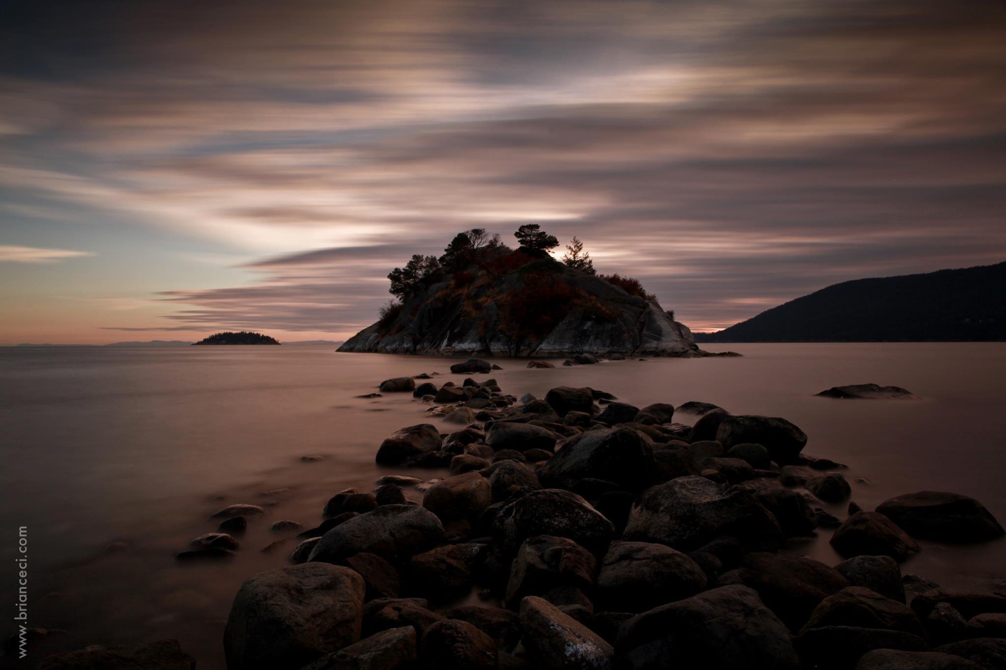 A rocky shoreline with smooth stones leads to a small island with trees, under a cloudy sky during dusk