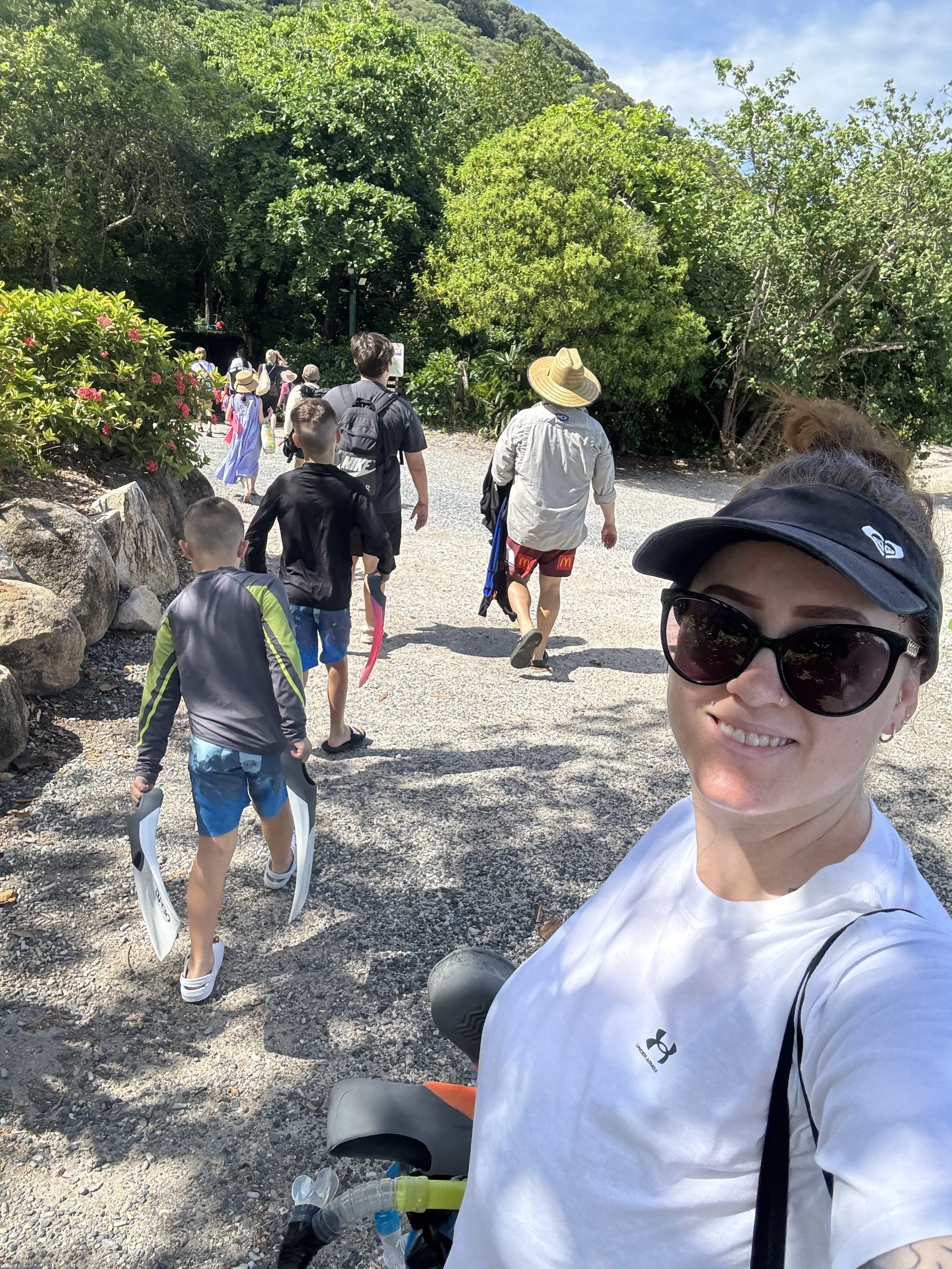 A smiling woman wearing sunglasses and a visor taking a selfie on a hiking trail with children and adults walking ahead under a bright, partly cloudy sky, surrounded by lush green trees and rocks.