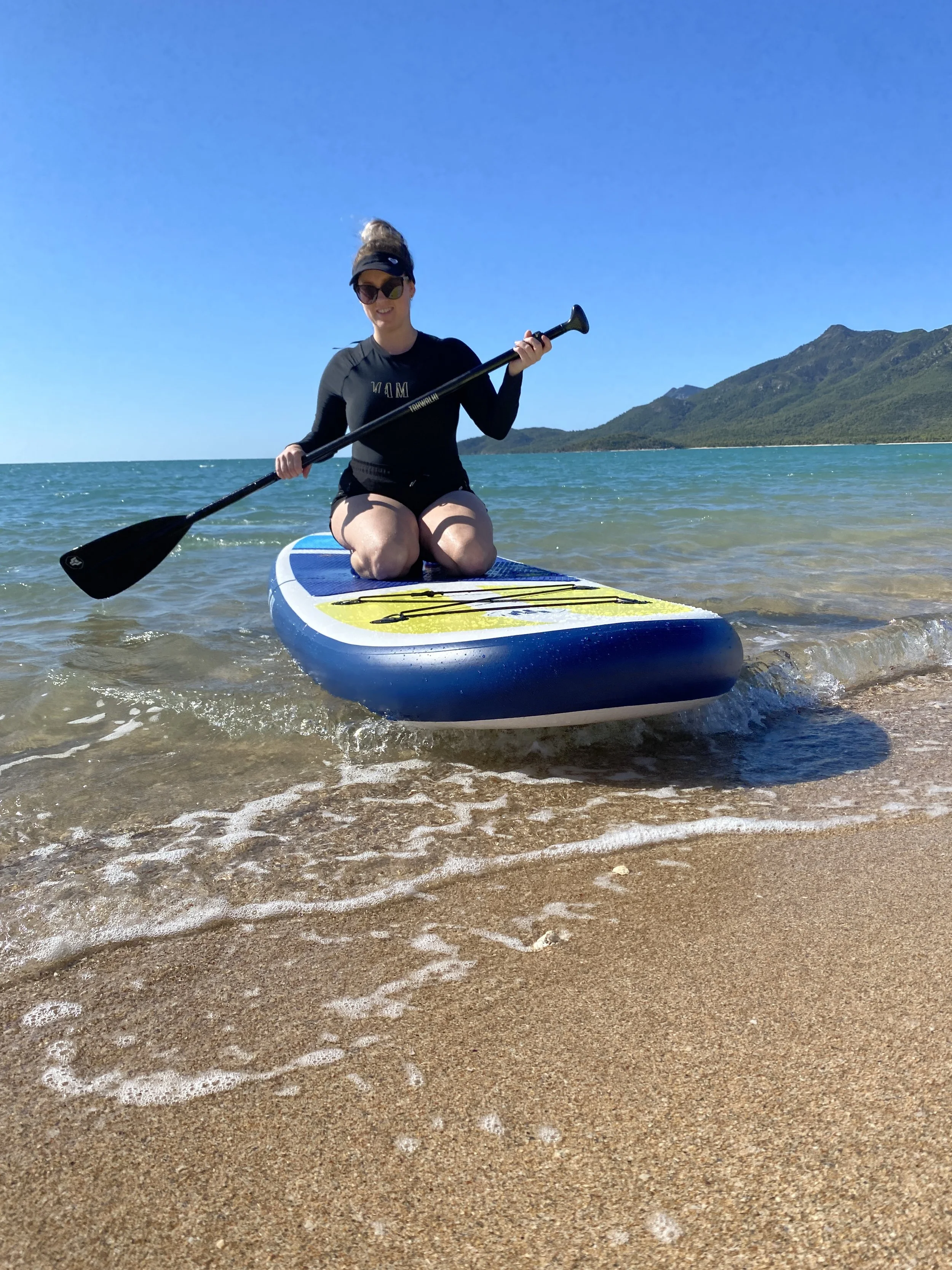 A woman kneeling on a paddleboard in shallow water near the sandy shore at the beach, holding a paddle and smiling, with mountains and a clear blue sky in the background.