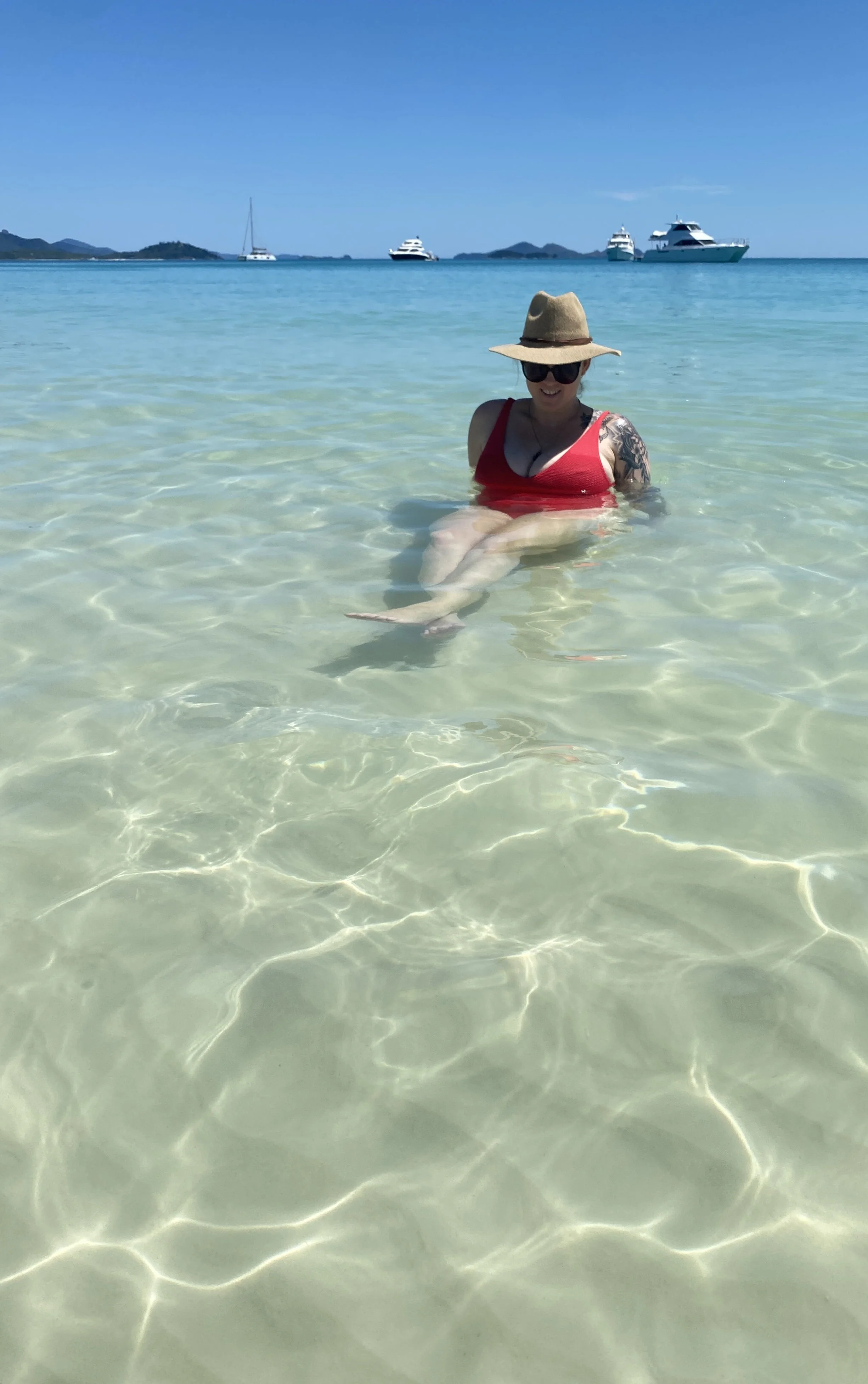 Woman in a red swimsuit and wide-brimmed hat sitting in clear, shallow ocean water with boats and islands in the background.