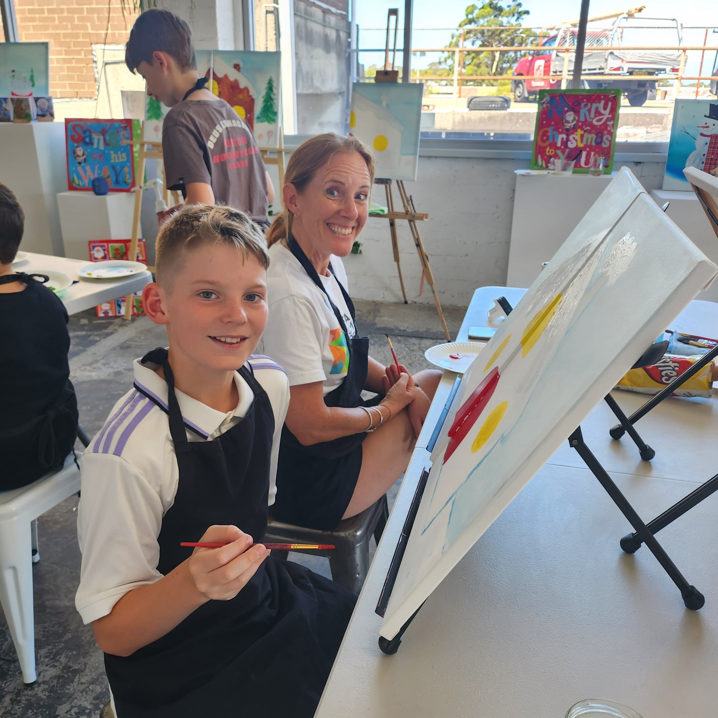 Mum and tween son attending Leigh Binskin Art's Adult and Child paint Date. Participants painting smiling at their canvases and enjoying themselves.