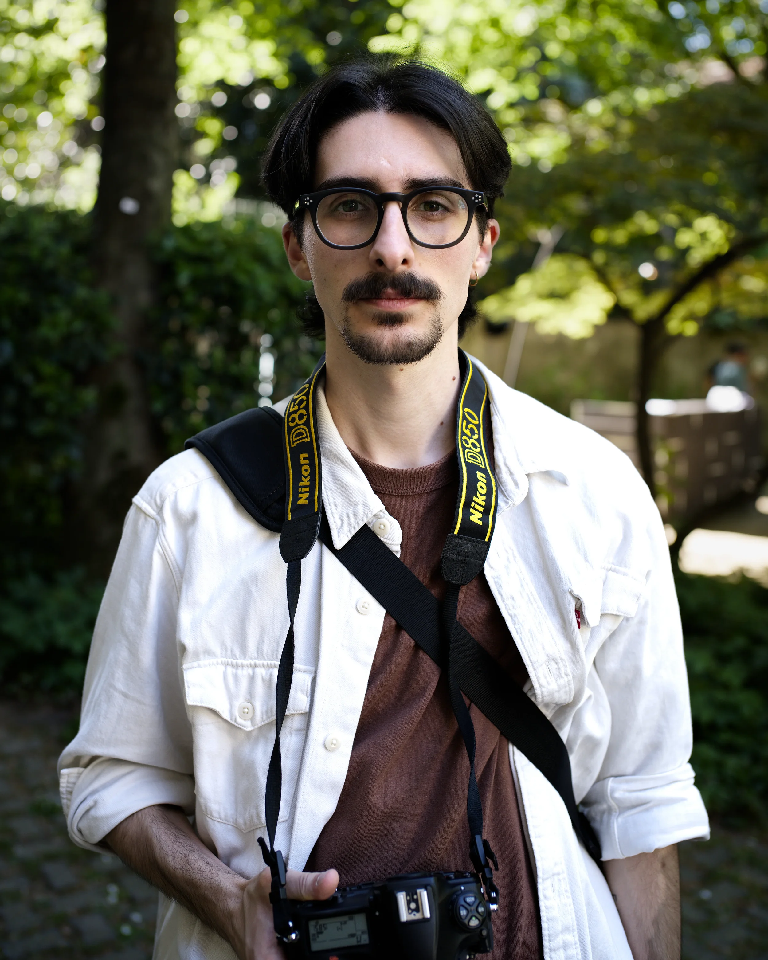 Un uomo con occhiali e baffi, con una fotocamera intorno al collo, in un parco verde.