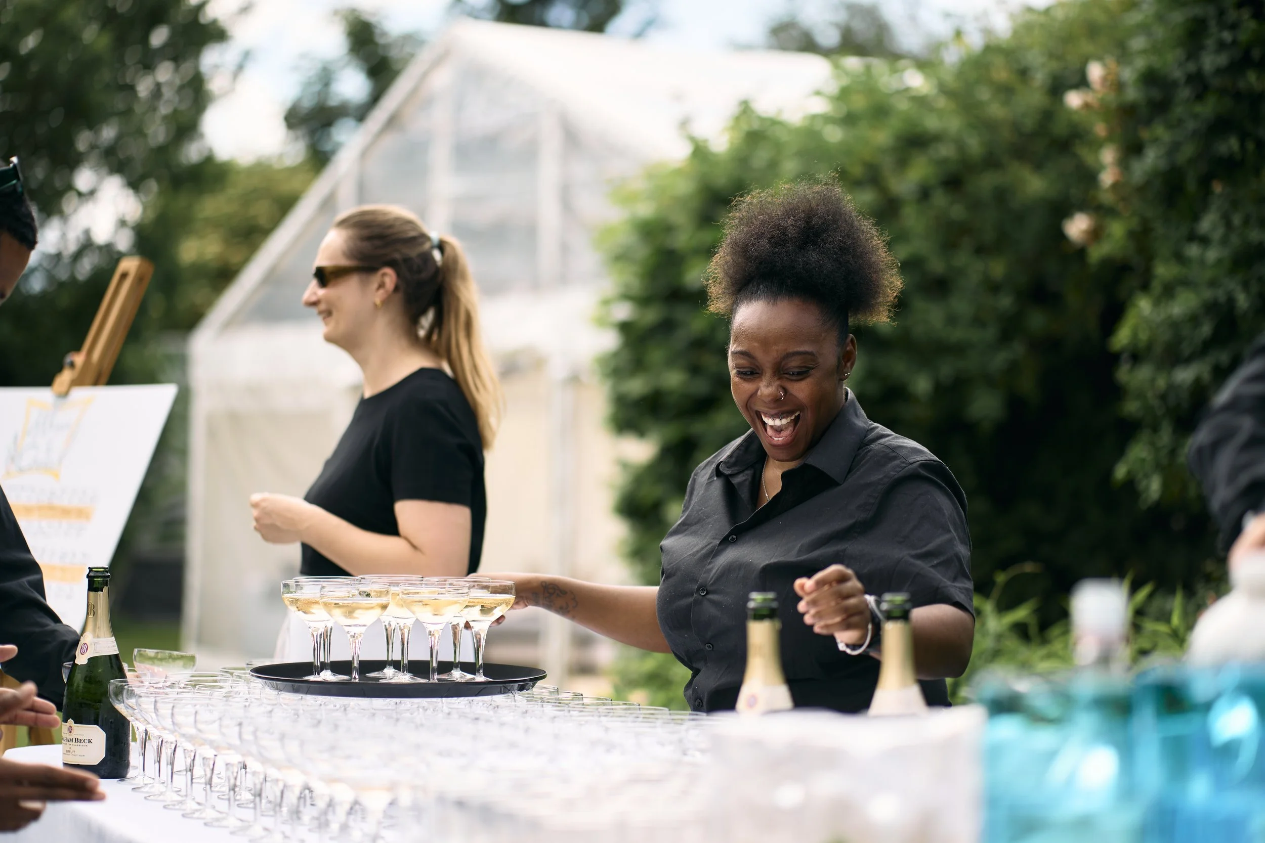 A group of women at an outdoor event, with one woman laughing as she serves drinks, and others smiling and engaging in conversation, with a table of champagne glasses and bottles.