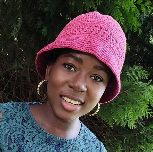 A woman of colour is facing the camera wearing the roo bucket hat in bright pink. The shape of the hat is bell like with a small short brim that sits close to the face. She is wearing hooped gold earings and a blue lace top. Green trees are behind.