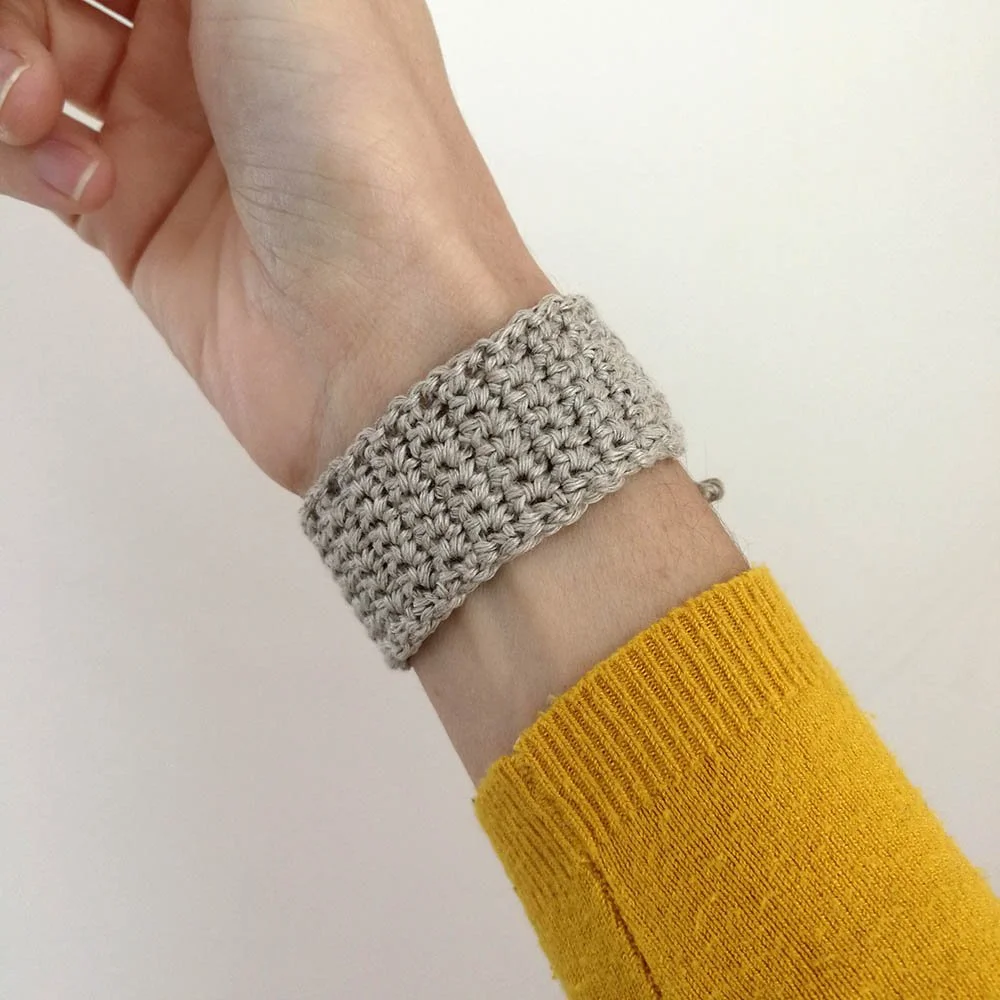 A right hand is being held up at an angle with the hand in the top left corner and the arm in the bottom right. The inside of the wrist is shown with the linen bracelet around it. The bracelet is light grey in colour and an inch wide.