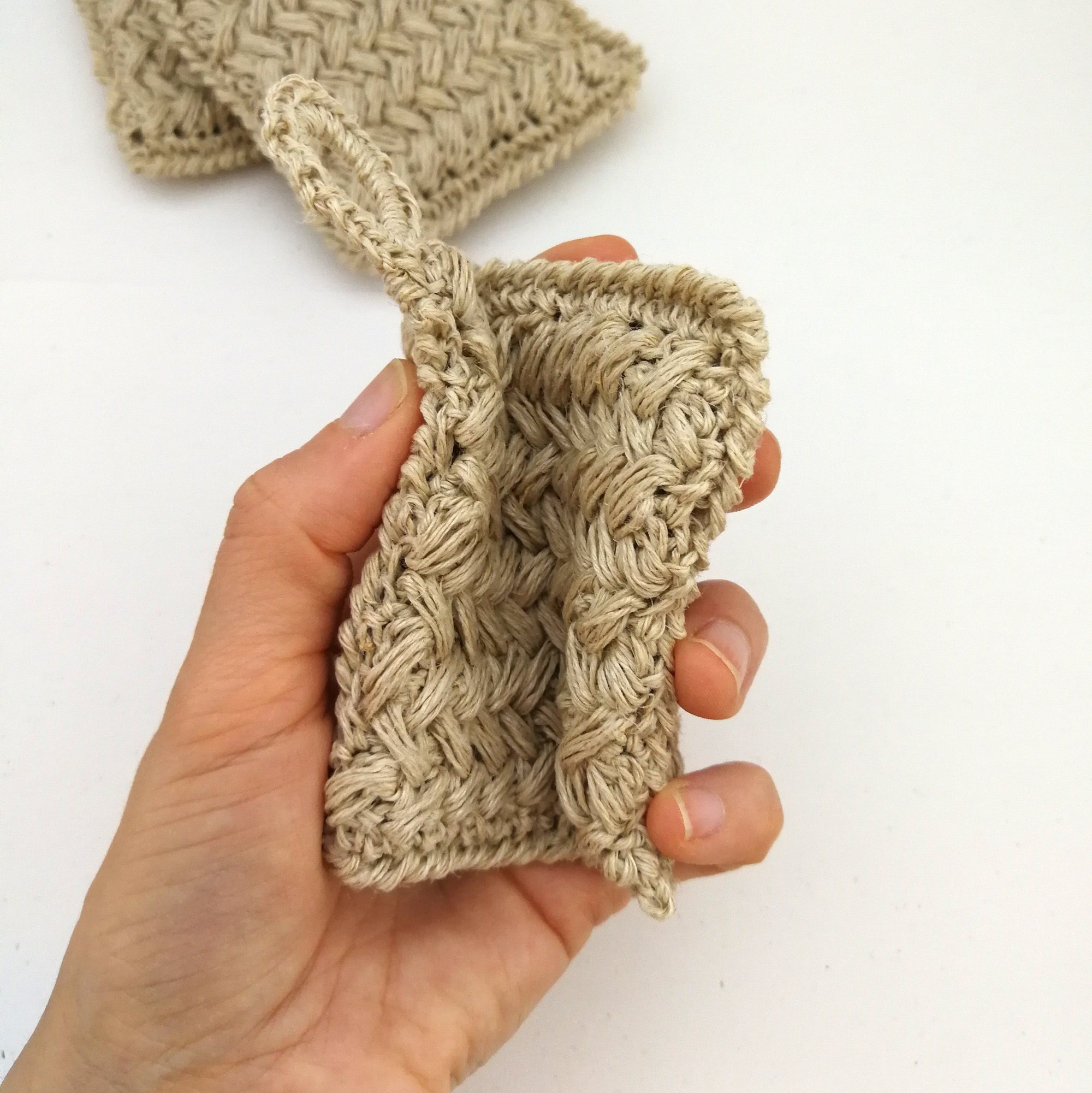 A left hand is holding 3 crochet kitchen scrubbies like a deck of cards. They have a braided type pattern. The sponge on the top has a small hanging loop on the top left.