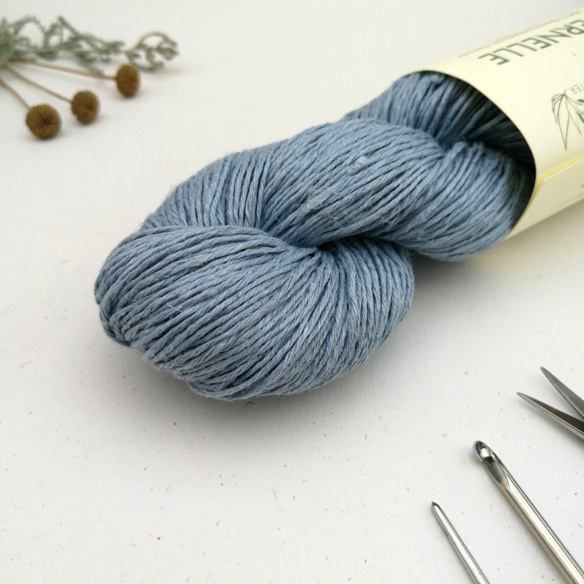 A skein of hemp yarn in bright baby blue is sat diagonally on a white background. A cream label is around its middle with green writing.