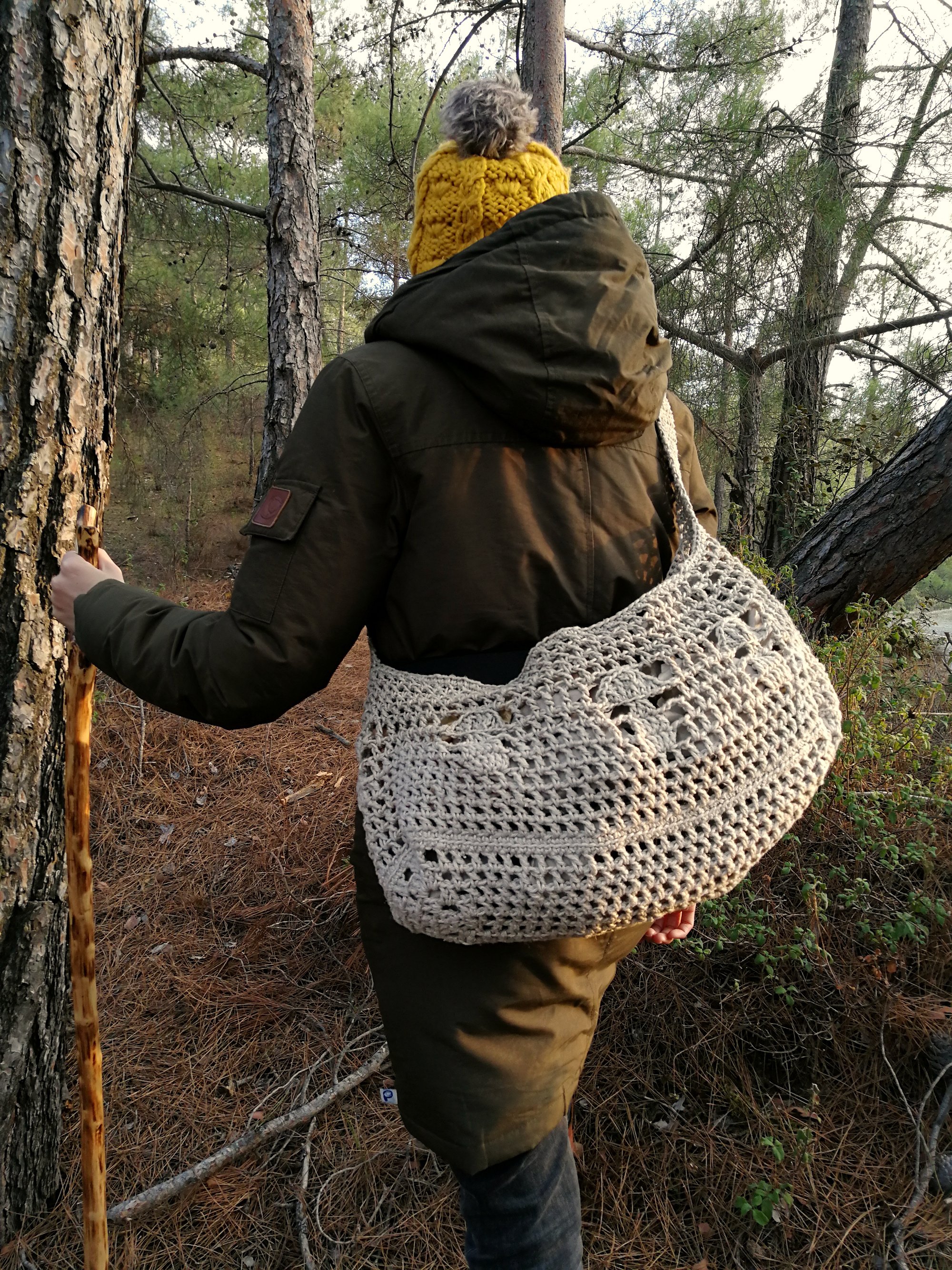 Tina is stood in the woods with the mushroom bag across her body with the bulk at her back. 3 crocheted mushrooms create a line across the middle of the bag.