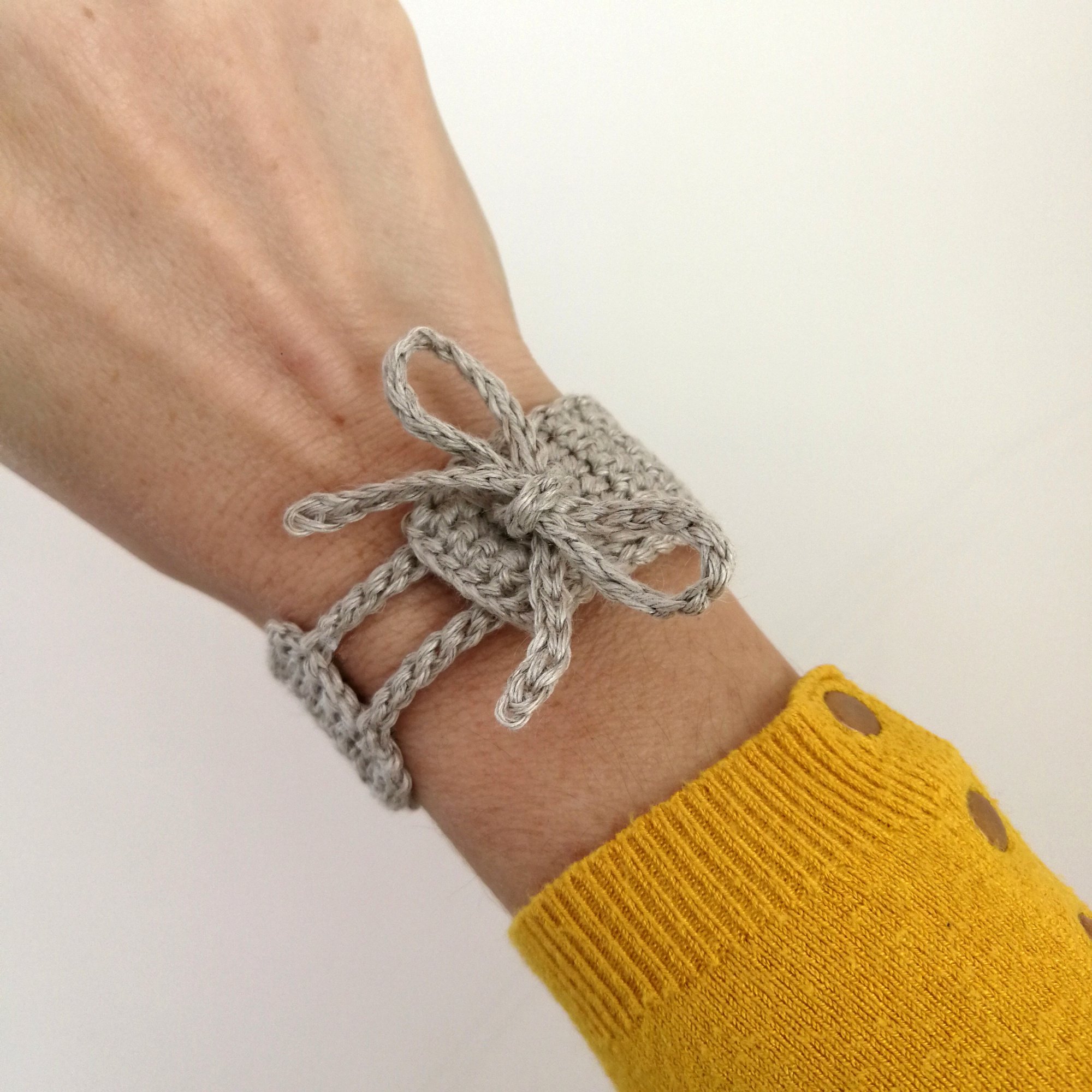 A close up photo of a right forearm wearing the linen bracelet which is light grey in colour and has a bow on the top. above the wrist is a yellow jumper with silver buttons down one side.