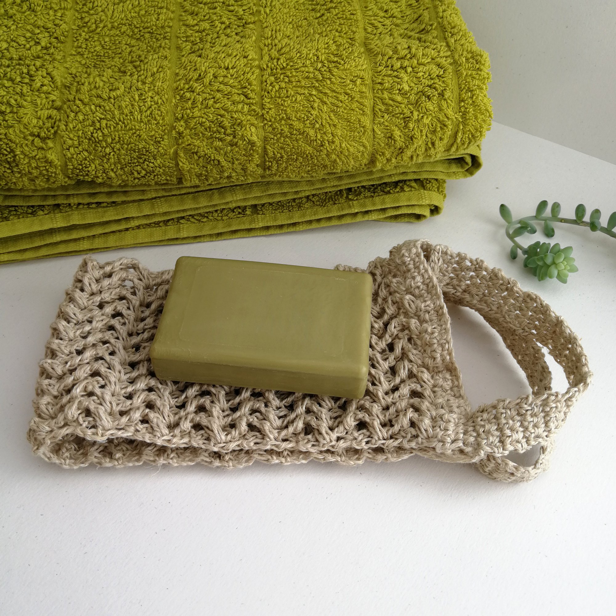 The back scrubber is folded in half & placed on a white table. The two sides are sat aside from each other & have a handle on each side. A green bar of soap is on the left and a green towel is behind.