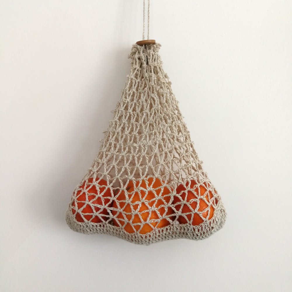 A natural produce bag is hanging from a hook against an off white wall. It has a V shaped mesh pattern and 3 mandarins inside it, side by side. A small, wooden button closes the bag at the top.