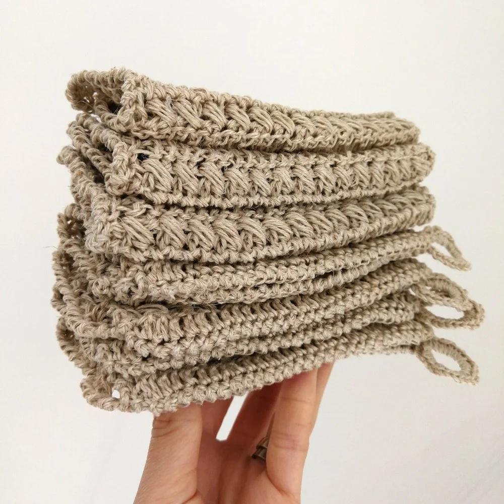 A white left hand is holding up 6 crocheted dishcloths on the tips of its fingers. The dishcloths are folded in half. Some have small hanging loops on the corners and some do not. They are all neutral coloured and the background is white.