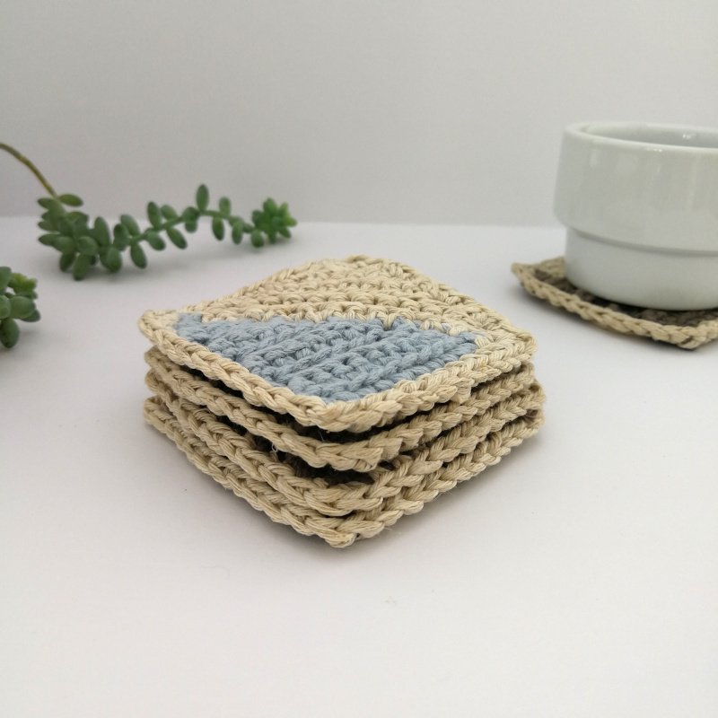 The coasters are in a stack with one of the corners facing the lens. In the background there is another coaster on the right with a white mug on it and there is a plant coming in from the left.