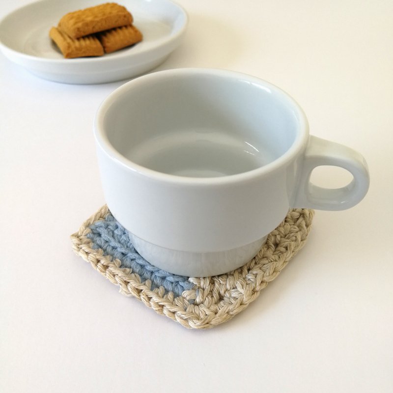 The baby blue square coaster is positioned in a diamond shape at the front of the photo. It has a small white mug on it and there is a small white plate of biscuits in the background.