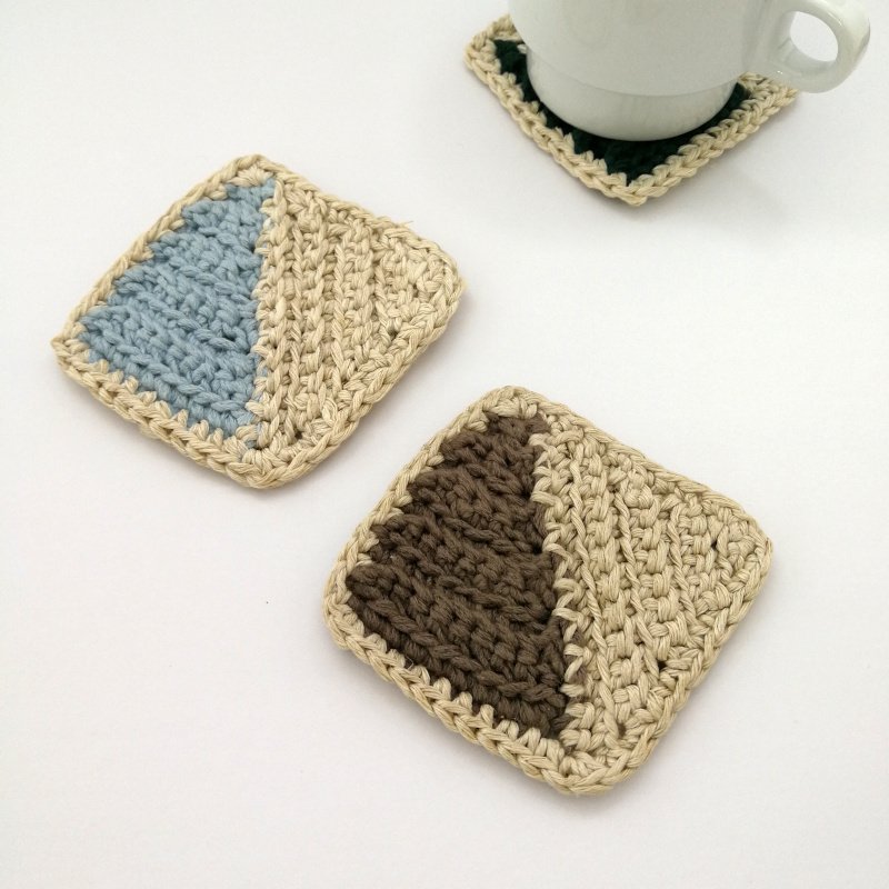 Two square coasters are sat on a white table with another coaster in the background with a white cup on it. Each coaster is made of two different coloured triangles and a border.