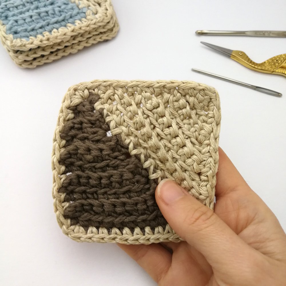 A right hand is holding the square coaster with the taupe coloured triangle. A small stack of coasters is in the top left corner with some crochet notions in the top right.