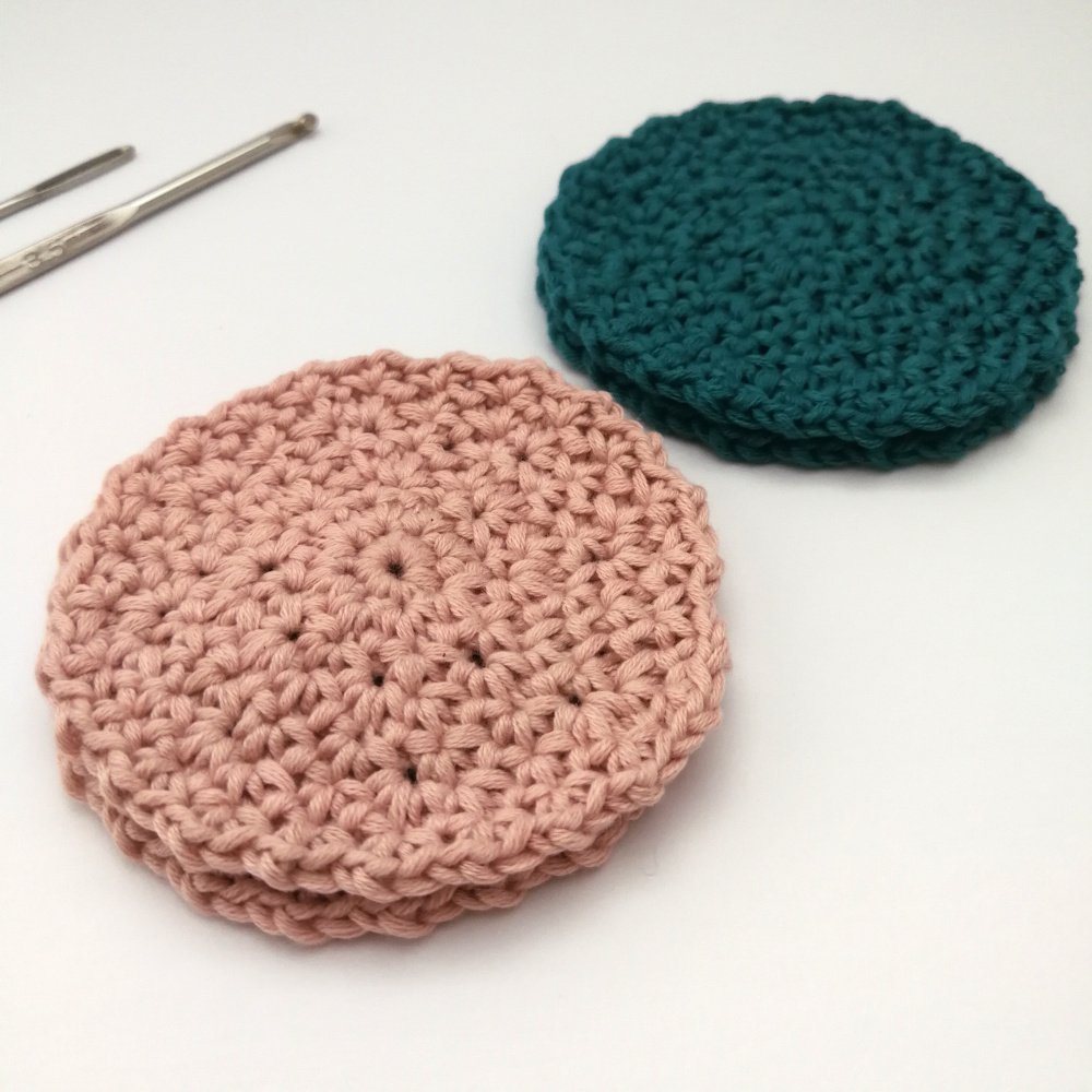 Two pastel pink face scrubbies are stacked and sat on a white table to the left of the photo and two more pastel teal face scrubbies are stacked behind more towards the right.