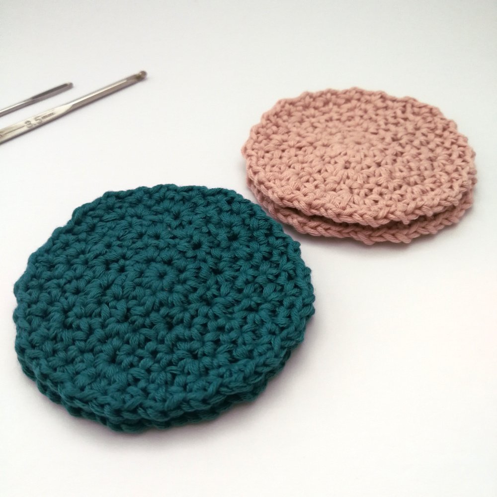 Two teal face scrubbies are stacked and sat on a white table to the left of the photo and two more pastel pink face scrubbies are stacked behind more towards the right.