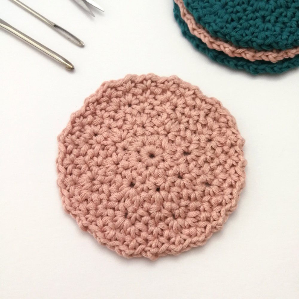 A birds eye view of a pastel pink face scrubbie. The very centre has a small hole and the rest of the scrubbie is an even textured of stitches with no clear beginning or end and an uneven edge.