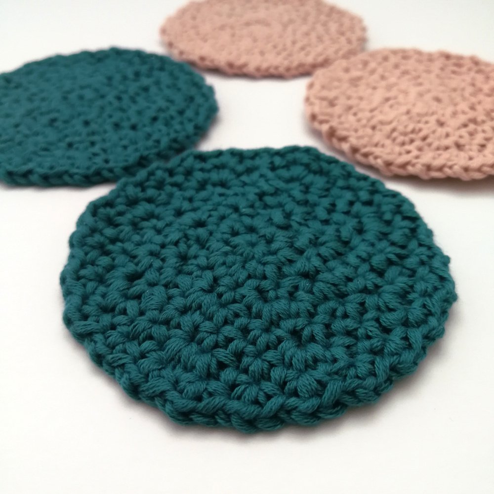 Four face scrubbies are sat flat on a white table. They are arranged in a cross shape with the south and west scrubbies being teal and the north and east scrubbies being pastel pink.