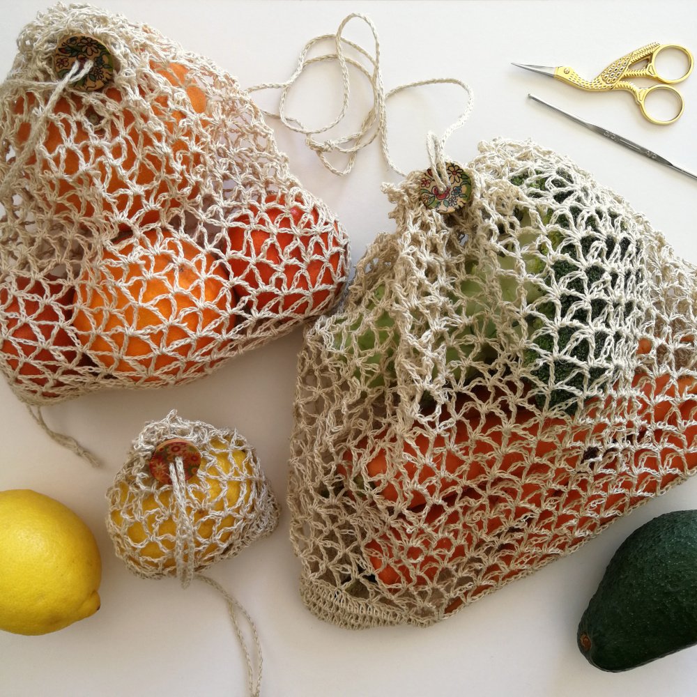 Three produce bags of different sizes made in natural hemp yarn are sat on an of white surface. They all have a V shaped mesh pattern an are filled with colourful fruit and veg.