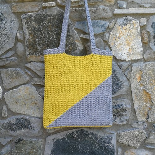 A person holding the crochet tote bag made from this pattern is stood facing left with the bag over their shoulder. The bag is mostly yellow with a grey triangle in the bottom right corner.