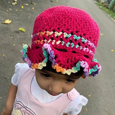 A toddler in a baby pink dress and white t-shirt is wearing a pink bucket hat with ruffles as a brim. The edge of the ruffles is multicoloured and there are a few multicoloured stripes around the body.