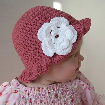 A baby wearing a pink top with dark prink flowers is facing to the right and wearing a dark pink crocheted sun hat. The hat has a large white flower stitch to the side and a ruffled brim.