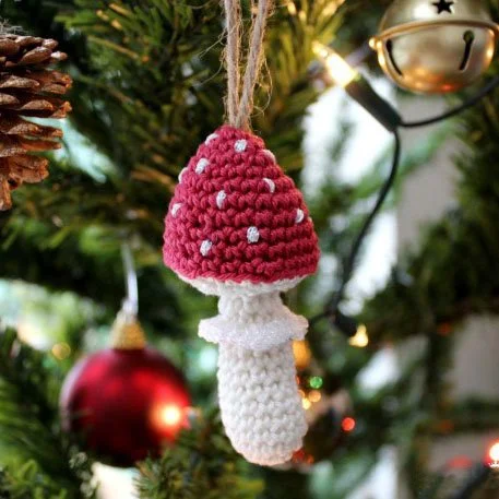 A close up photo of a cute little crocheted mushroom with a white body and a red pointed top with white polka dots hanging from a Christmas tree with some natural coloured string.