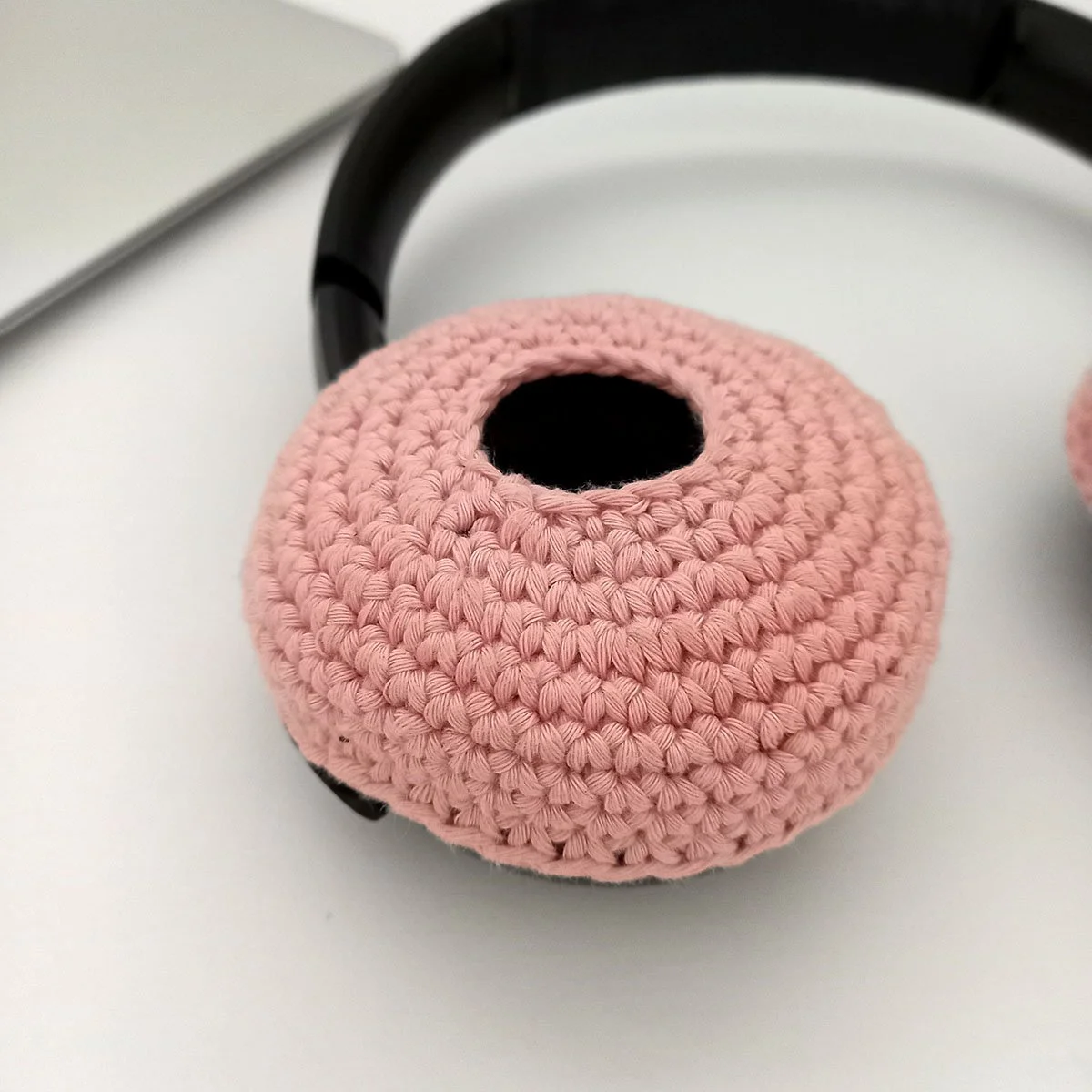 A side angle view of the left side ear piece with the pastel pink cover ontop. The single crochet stitches have a clear definition. The open centre looks like a black hole. The black head band is visible in the background along with the silver laptop