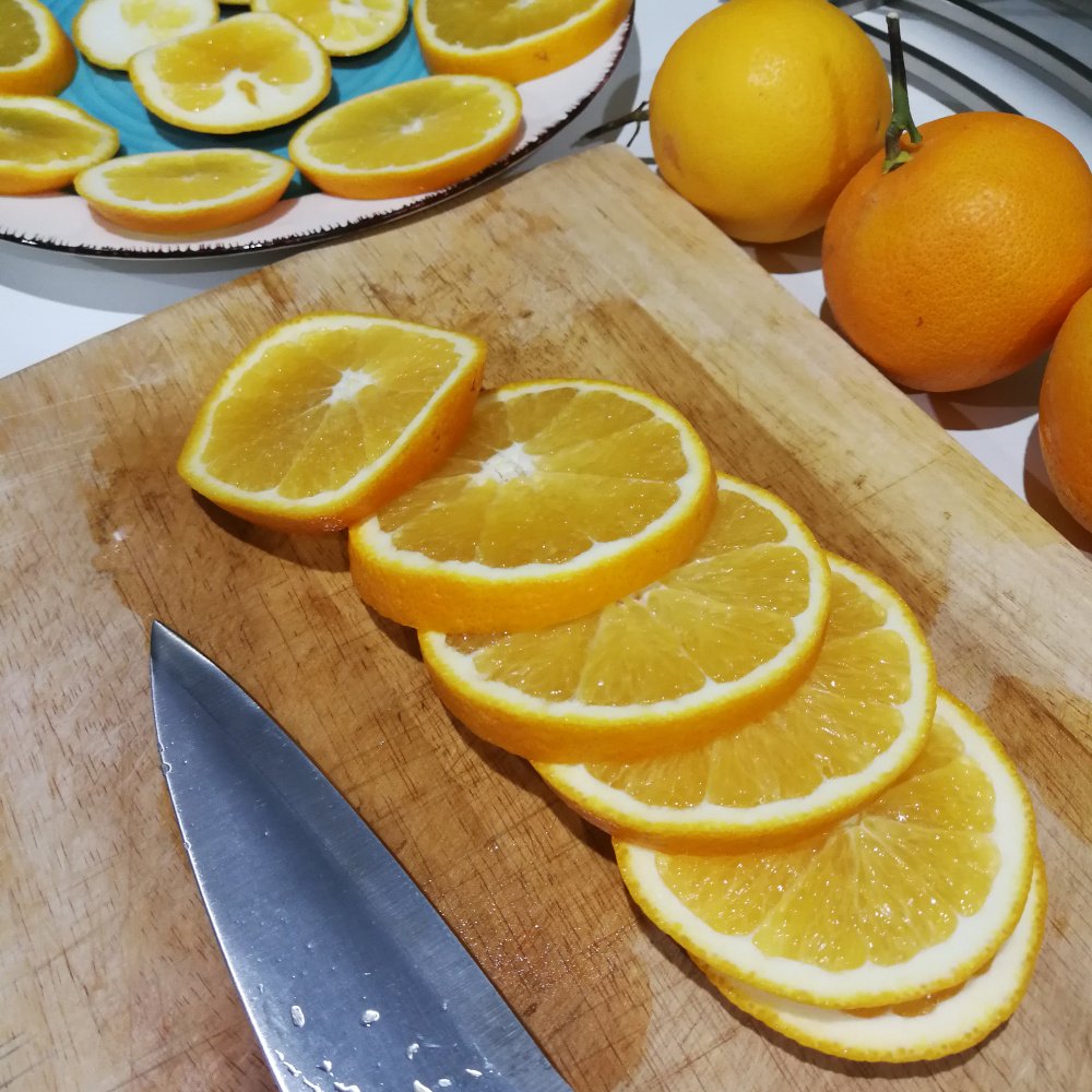 Six slices of orange are layered in a row on a wooden chopping board. A large silver knife is parallel. There is a plate in the background with more slices on it and 3 full oranges to the right.