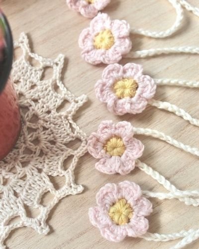 A portrait image of 5 small crocheted flowers aligned from top to bottom. They have a yellow centre and pink petals with a white crocheted chain attached. They are sat on a pale wooden surface and on the left is a crocheted doily with a plant pot.