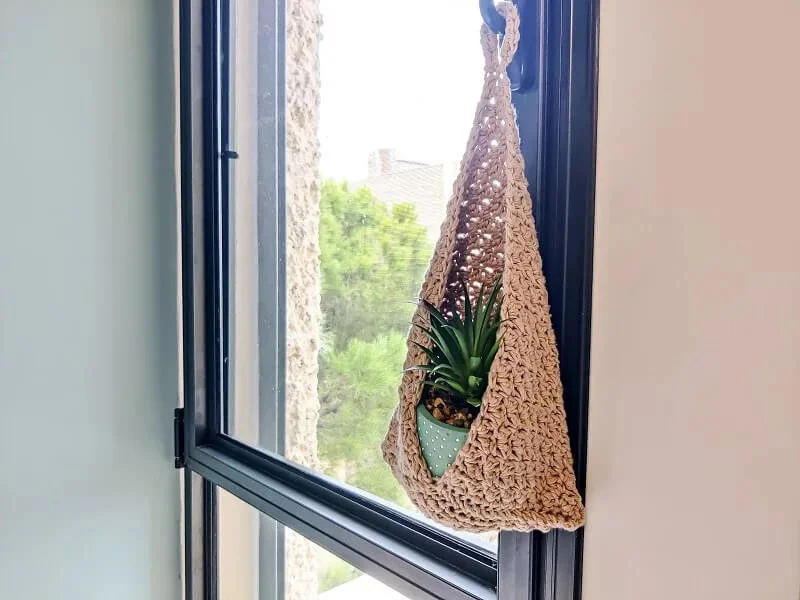 A beige, triangular shaped basket is hanging from the handle of a dark grey window frame. There is a green spikey plant in a light teal pot inside in the basket.