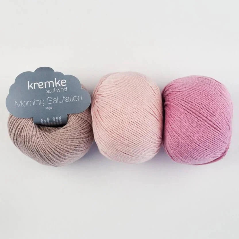 A birds eye view of three soft looking balls of yarn in taupe, pale pink and pink. Taupe has a grey yarn label in the middle with white writing that reads "Kremke". The other two are on their side.  The background is off grey.