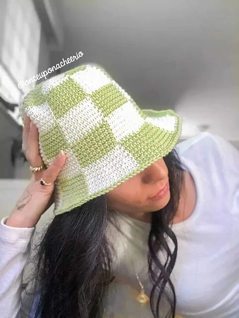 A person of colour with long back wavy hair is resting their left elbow on a surface with their head in their hand. They are wearing a green and white bucket hat in alternating coloured squares.