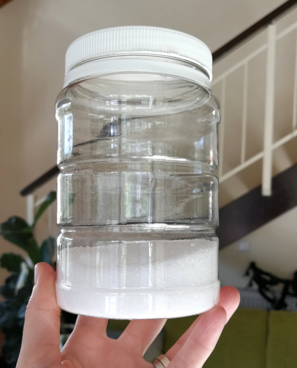 A left hand is holding up a plastic container filled nearly 1/3 of the way up with citric acid. The container is clear and the lid and the contents are bright white. The background is a staircase and some houseplants.