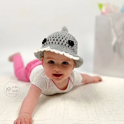 A baby in a white spotted top and pink trousers is lying on their front on a cream surface wearing a grey shark bucket hat with black eyes, a fin at the top and a white toothed brim. The baby is smiling at the camera with their arms stretched out.