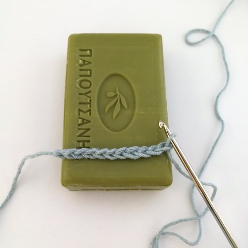 A green bar of soap laid in a portrait position with the foundation chain made in baby blue yarn laid across it. The silver crochet hook is still attached to the working loop and the background is white.