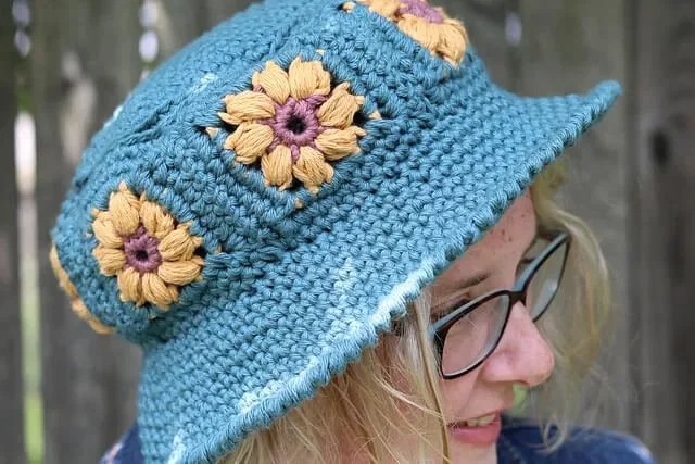 A close up photo of a white person with blonde bob length hair facing to the right. They are wearing the garden bucket hat which is turquoise with yellow and pink flowers around the body.
