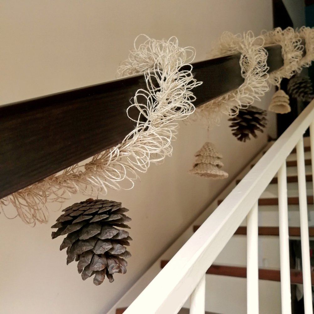 A dark wooden banister with white metal railings has the natural coloured eco friendly tinsel wrapped around it. The photo is taken from the bottom of the steps looking up along the banister. It also has pine cones & mini xmas trees hanging from it.