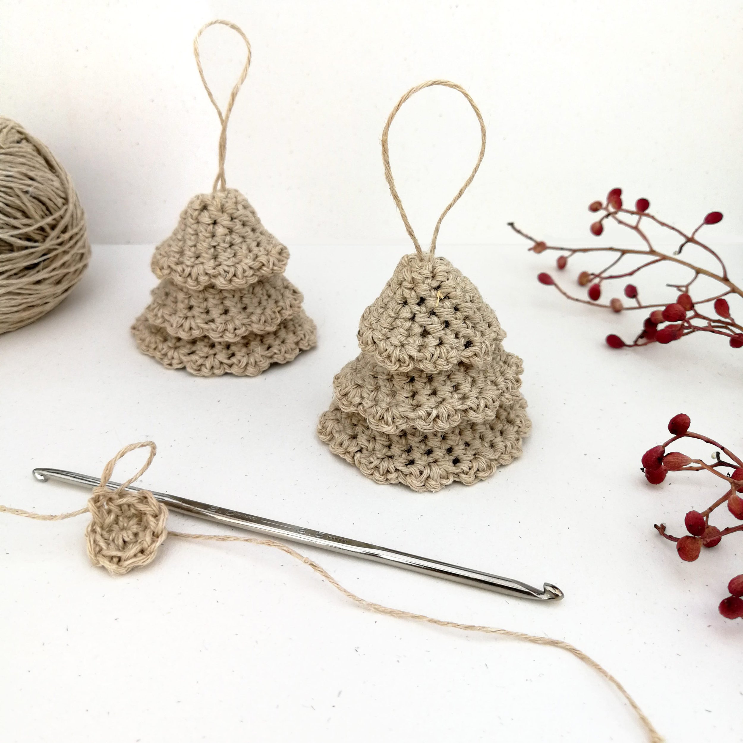 Two crocheted Christmas trees stand against a white background. They have three skirts stacked on top of each other to make the tree & a loop at the top. There are some small red berries along the right hand side & a ball of yarn in the top left.