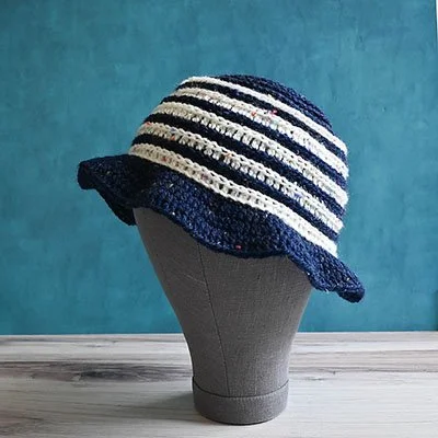 A navy and white striped bucket hard with a wavy brim is placed on a grey mannequin head. The head is sat on a grey wooden surface and the wall behind is turquoise.