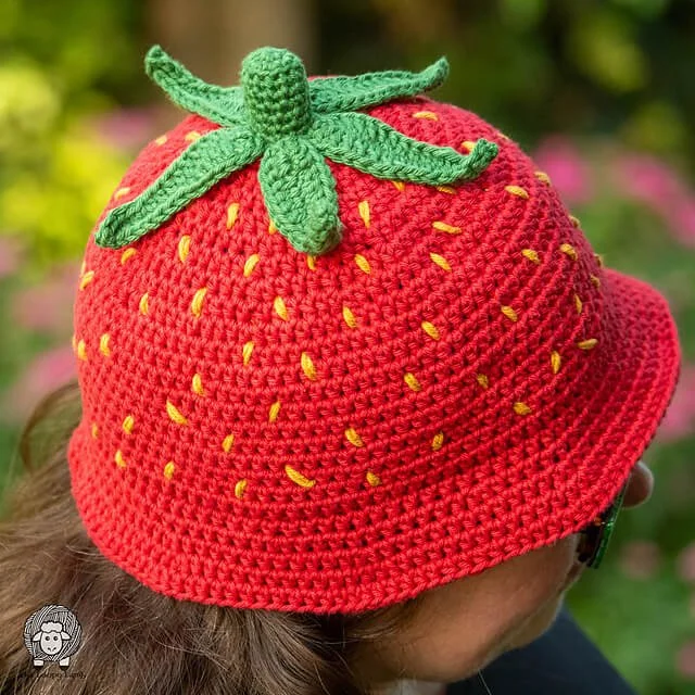 A close up photo of a white person wearing the hat. They are facing to the right with their face obscured by the hat. The hat is bright red with small yellow flecks all over and a green stem and 5 leaves on top. It has a small brim.