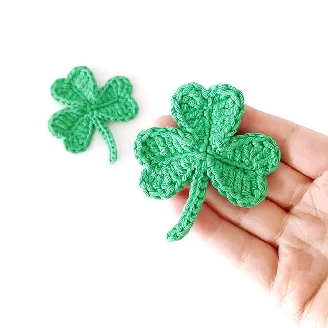 A birds eye view of a right hand holding a crocheted shamrock on the end of their fingers. The background is white and below the hand there is another shamrock laid on the white surface. They are both bright green.