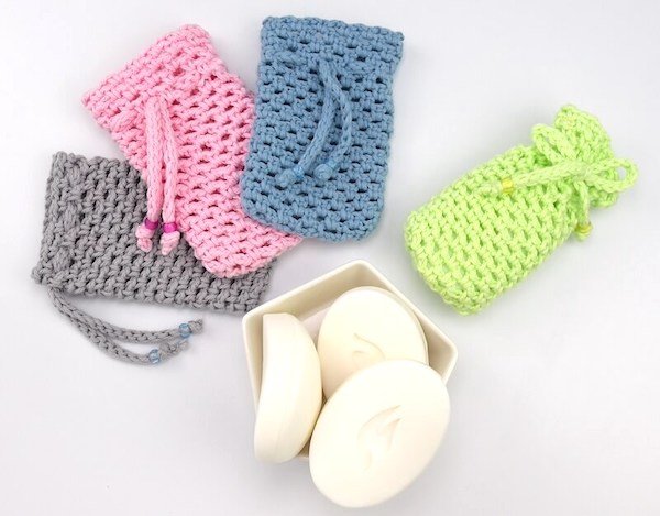 Four different coloured crochet soap savers are laid out in a fan shape on a white background. From left to right they are grey, pink, blue and lime green. There is a hexagonal shaped white container with no lid with 3 bars of soap inside.