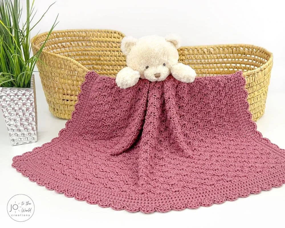 A dusty pink baby blanket is draped over a woven baby basket. There is a white teddy bear peering out over the middle of the baskets edge and the blanket towards the camera. The blanket is sprawled out across the floor in front.