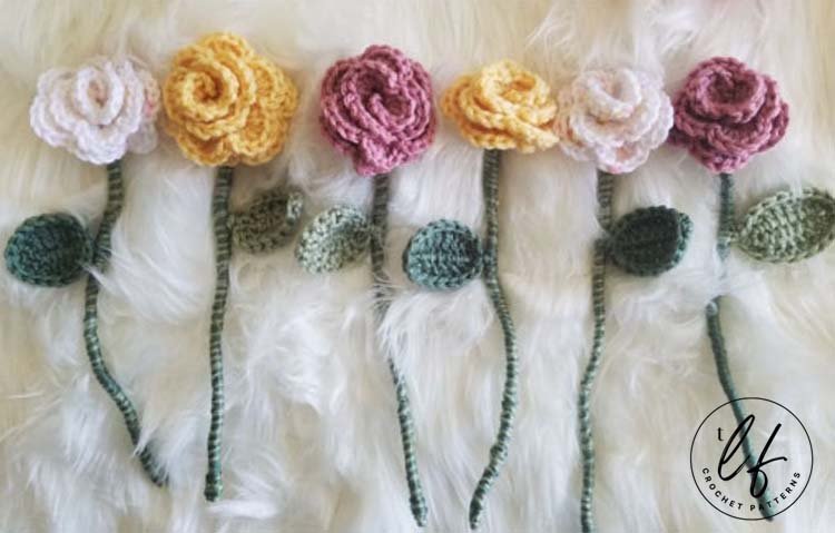 A rectangular photo of 6 crocheted roses sat on a white furry surface. The roses are solid colours of white, yellow and pink with green stems and 1 green leaf each.