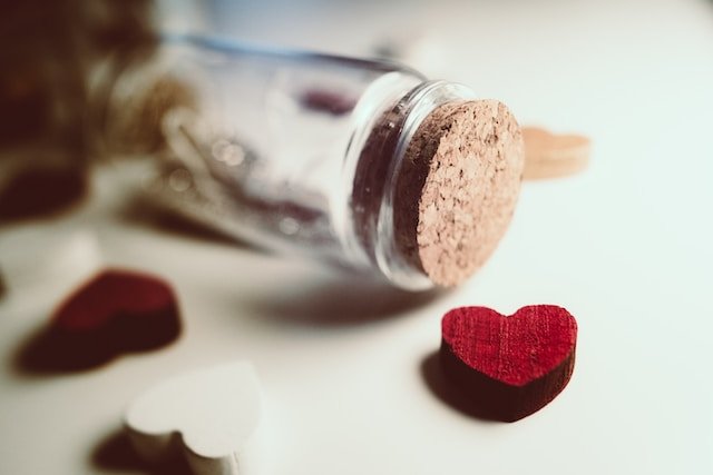 A photo of a small clear glass jar with a cork lid laying on a white surface with the lid pointing to the right.  There is a red wooden heart in front of it and some others scattered around that are solid colours of white or red.